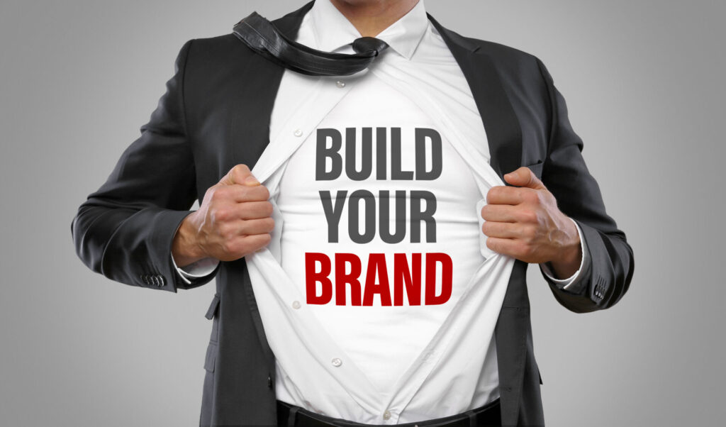 A professional man in a suit pulls open his shirt to reveal the words “Build Your Brand,” symbolizing a Realtor developing a strong personal brand in real estate.